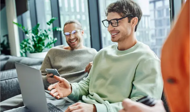 Two men sitting on a couch, one typing on a laptop while the other looks on attentively.