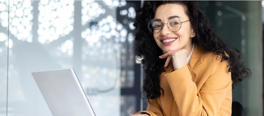 A smiling woman wearing glasses is working on her laptop, exuding a sense of joy and productivity.