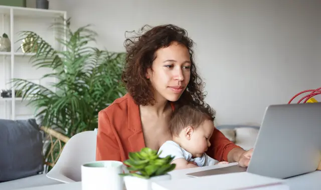 A woman multitasking by holding her baby and using a laptop.