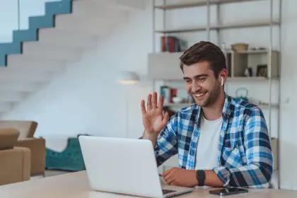 A man waves while sitting at a table with his laptop.