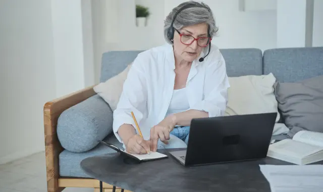 Elderly woman sitting on couch with laptop and headphones.