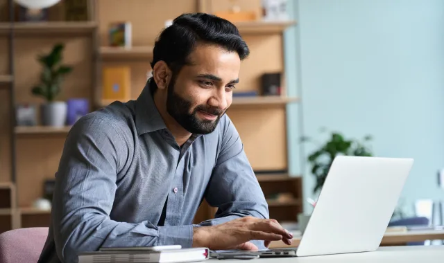 A bearded man in a shirt focused on his laptop screen.