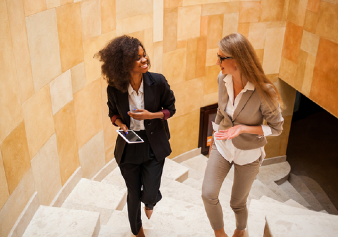 Two businesswomen confidently ascend a staircase, engaged in conversation and dressed in professional attire.