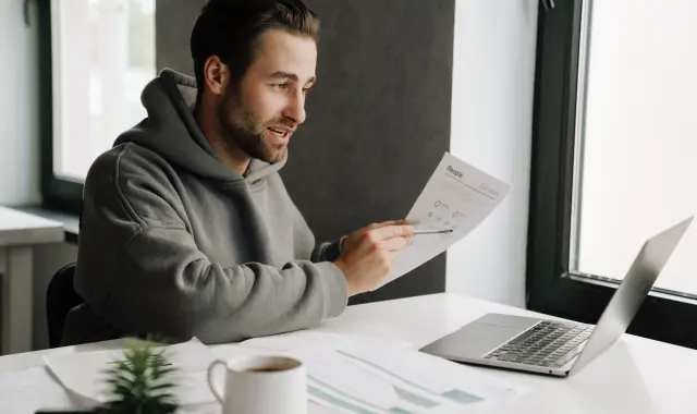 A man in a hoodie focused on a document on his laptop screen.